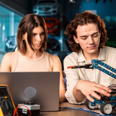 Young man and woman in protective glasses doing experiments in robotics in a laboratory using a computer. Robot and tools on the table
